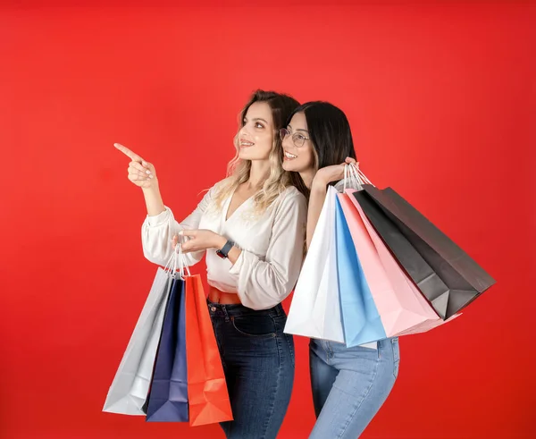 Two best friends carrying shopping bags. Portrait of young caucasian lady, cute, nice, charming, long haired teenage girl carrying new purchase. Pointing copy space. Isolated warm red background.