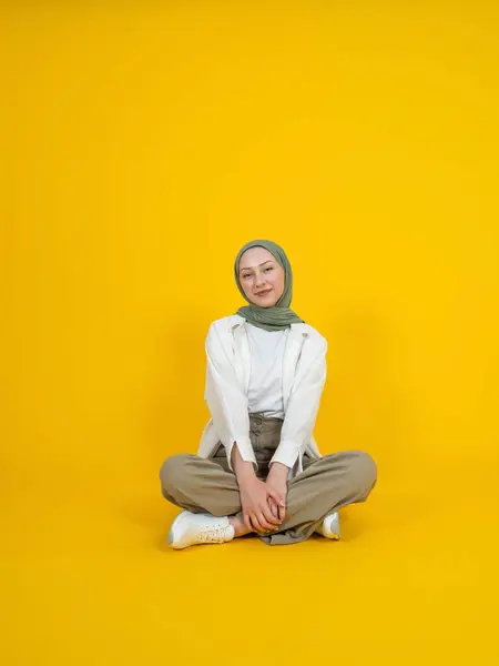 Caucasian muslim woman, full body young smiling calm caucasian muslim woman wear hijab. Sit floor hold legs. Yellow studio background copy space. Looking camera. Peaceful islam religious lifestyle.