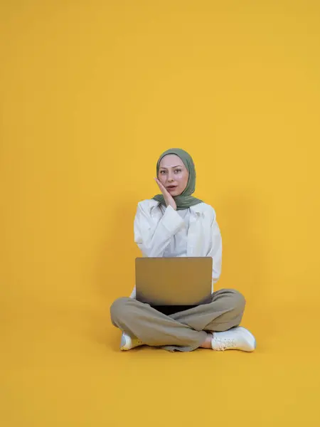 Shocked muslim woman in hijab, full body length caucasian shocked muslim woman in hijab sit floor legs crossed with laptop, keep mouth open put hand on cheek. Isolated yellow background.