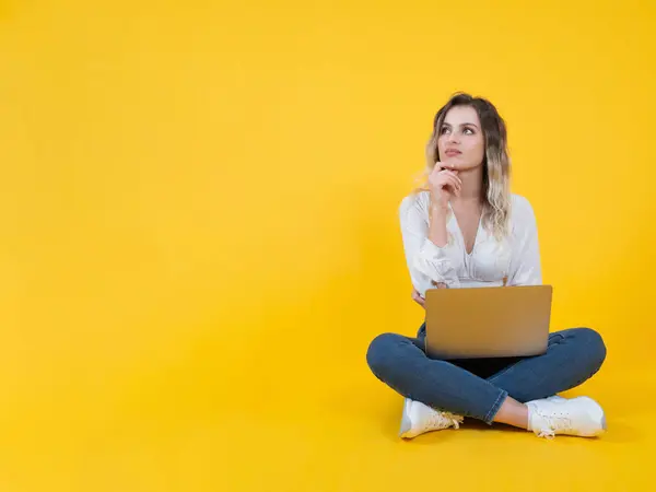 Pensive woman, full body young blonde pensive woman. Sit floor isolated yellow background hold use laptop. Working online, freelance. Looking aside put hand prop up chin. People lifestyle concept.