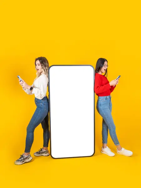 Mobile application advertisement, full body young two women leaning big mobile phone with empty blank screen mock up. Isolated yellow background. Copy space. Using smartphone, looking display.