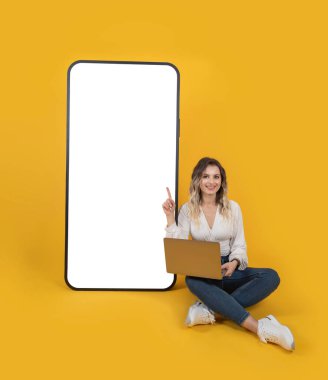 Büyük boş ekran telefonu, tüm vücudu oturan sarışın kadın dizüstü bilgisayarı kullan ve büyük boş ekranlı telefonu işaret et. Çok büyük bir akıllı telefon modeli. Yeni uygulama, teklif, dijital reklam gösterimi. 