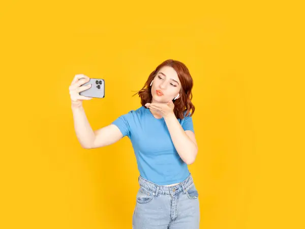 Taking selfie blowing air kiss, portrait of pretty young red bob hair caucasian woman taking selfie blowing air kiss. Standing over yellow studio background. Wear casual blue shirt and jeans.