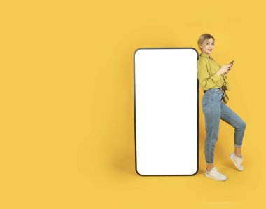 Mobil uygulamayı kullanarak, uzun boylu sarışın kız beyaz dokunmatik ekranlı büyük, boş bir ekran telefona yaslanıyor. Akıllı telefon kullanarak, kameraya bakarak. Sarı arkaplan, boşluğu kopyala.