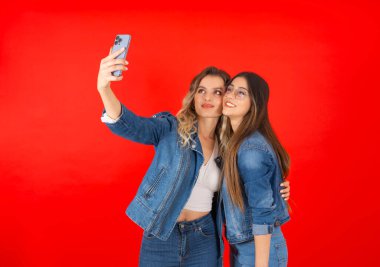 Selfie çekmek, iki genç bayan en iyi arkadaşın selfie çekerken portresi. İnternetten, sosyal medyadan hava öpücüğü yollamak. Kot ceket ve kot pantolon giy. İzole kırmızı arka plan. Boşluğu kopyala.