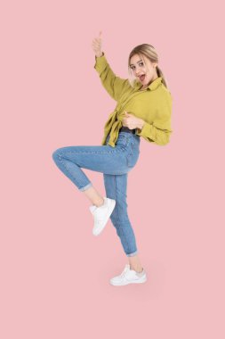 20 'li yaşlarda dans eden, kutlama yapan, baş parmağını gösteren beyaz bir sarışının tam vücut fotoğrafı. Sarı gömlek, kot pantolon ve beyaz spor ayakkabı giy. İzole pembe arka plan. Boşluğu kopyala.