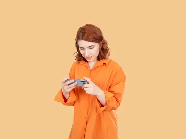 Play online mobile game, photo of focused young red bob hair woman play online mobile game. Posing isolated over pastel orange background. Hold use mobile phone, watching video on smartphone.