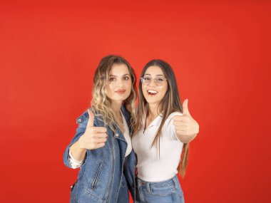 Başparmaklar yukarı, genç, güzel, beyaz, harika iki kadının portresi kırmızı stüdyonun arka planında baş parmak hareketi gösteriyor. Sıradan kot pantolon giyin. Önerin, konsept teklif et.