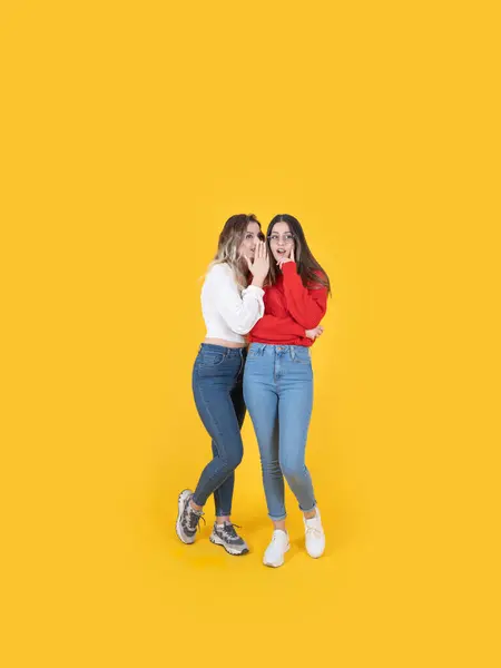 Whispering secrets, full body length caucasian best friends standing yellow studio background. Share gossip together, spread rumors. Wear casual dress denim jean. Mouth to ear words concept.