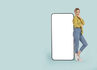 20 'li yaşlarda, sarışın, beyaz bir kız büyük bir cep telefonuna yaslanıyor ve boş beyaz dokunmatik ekranla fotokopi alanını işaret ediyor. Uygulama tanıtımı.