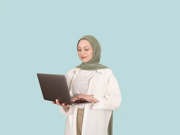 Woman in hijab using laptop, portrait of 20s caucasian attractive confident woman in hijab using laptop. Wear casual abaya holding carrying typing computer pc notebook. Isolated blue background.