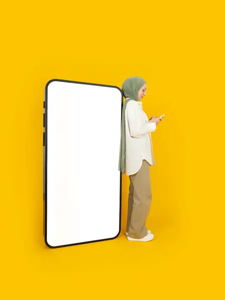 Full body caucasian muslim 20s woman in hijab leaning back huge big empty blank white screen mobile phone mock up over isolated yellow studio background. Islam religious lifestyle concept image.
