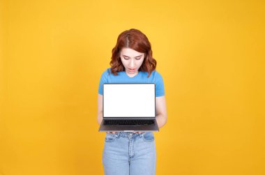 Kızıl saçlı, beyaz saçlı, elinde boş bir dizüstü bilgisayar tutan genç bir kadın. Sarı stüdyo arkaplanının üzerinde dur, kopyalama alanı. Halk yaşam tarzı, tavsiye edilen web sitesi, yeni uygulama kavramı.