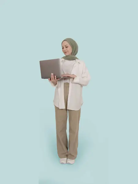 Muslim woman in hijab posing with laptop, full body length positive smiling caucasian islamic lady standing over blue background using computer for work, online shopping, browsing internet.