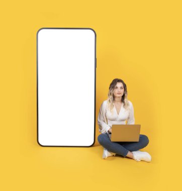 Büyük cep telefonu maketi, tüm vücut uzunluğunda güzel sarışın kadın dizüstü bilgisayarı kullan büyük cep telefonu modelinin yanında oturan kamera kullan. Boş büyük beyaz dokunmatik ekran. Reklam kavramı.