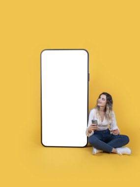 Çevrimiçi alışveriş uygulaması, tam vücut ölçülerinde sarışın beyaz kadın büyük boş ekran cep telefonunun yanında oturuyor. Elinde kredi kartı ve akıllı telefon görünümlü beyaz dokunmatik ekran fikri var..