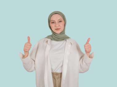 Baş parmaklar havada, 20 'li yaşlarda beyaz Müslüman bir kadının portresi onaylıyor. Kendine güvenen genç girişimci, iş kadını ya da mavi arka planda poz veren öğrenci. Boşluğu kopyala.