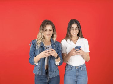 Cep telefonu kullanan iki kadın, genç beyaz bir kadının fotoğrafı cep telefonu kullanan iki kadın dokunmatik ekranda gülümsüyor. Kırmızı stüdyonun arka planında dikiliyoruz. Kot pantolon giy, yaşam tarzı konsepti.