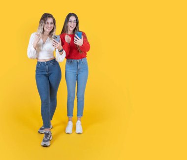 İki kadın kazanan jest, tüm vücut görünümü, heyecan verici iki beyaz kadın kazanan jest. Cep telefonunu kullanıp gülen surat ifadesini ekrana yansıt. Sarı arkaplan, boşluğu kopyala. 