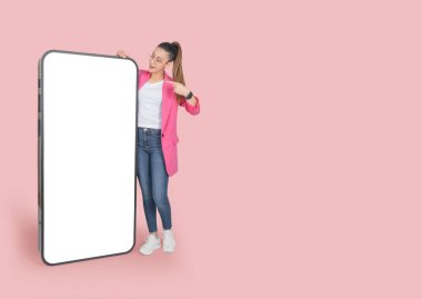 Kocaman bir cep telefonu modeli, koca vücutlu, beyaz tenli, koca bir cep telefonu modeli çizen bir kadın. Boş dokunmatik ekranı işaret ediyor. Pembe resmi ceket giyin, izole edilmiş, kopyalama alanı.