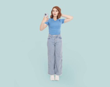 Kafası karışmış bir kadın, tüm vücudu beyaz, kafası karışmış genç bir kadın elinde modern akıllı telefon tutan kamerayla kafasını kaşıyan bir kadın. Açık mavi arka planda dikiliyorum. Şaşırmış yüz ifadesi. 