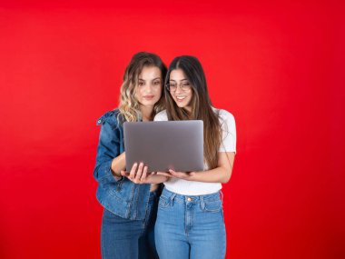 Dizüstü bilgisayar kullanan iki güzel beyaz kadın dizüstü bilgisayar kullanıyor. Ekrana bakmak, video izlemek, izole edilmiş kırmızı arka planda gülümseyen pozitif kızlar. Kot pantolon giy, kopyala.