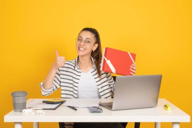 Hediye kutusunu tutan, çekici iş kadını ofis masasında oturan, elinde hediye kutusuyla mutlu bir gülümseme gösterisi yapan, baş parmağını kaldır. Sarı stüdyo arka plan, kopyalama alanı. Kariyer konsepti.