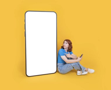 Mobil uygulama reklamı, tüm vücudu beyaz saçlı genç beyaz saçlı kadın kocaman bir telefonun yanında oturup boş beyaz dokunmatik ekranı işaret ediyor. Sarı arka plan. Boşluğu kopyala.