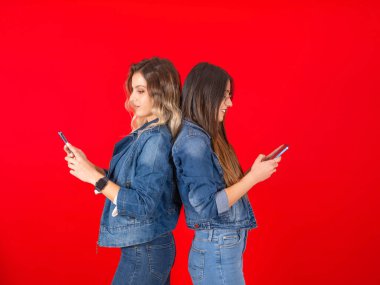Yan görüş genç iki beyaz kadın cep telefonu kullanıyor kot ceket ve pantolon giyiyorlar. Kırmızı stüdyonun arka planında arka arkaya akıllı telefon ekranı görünüyor. Boşluğu kopyala.