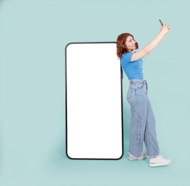 Telefon modeli yap, tüm vücudu gör genç kızıl saçlı beyaz kadın telefonun yanında duruyor. Kocaman, boş, dokunmatik akıllı telefon. Kız yakınlarda selfie çekiyor. Çevrimiçi reklam sunma kavramı