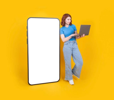 Mobil uygulamayı kullanarak, 20 'li yaşlarda kadın arkasını yaslıyor büyük akıllı telefon boş beyaz dokunmatik ekran tutuş dizüstü bilgisayarı ile taklit ediyor izole edilmiş sarı stüdyo arkaplanı üzerinden mobil uygulama kullanıyor..