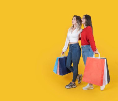 Full body length side view two beautiful fit caucasian 20s young women wear casual slim jeans and shirt carry many shopping bags walking looking aside copy space over isolated yellow studio background clipart