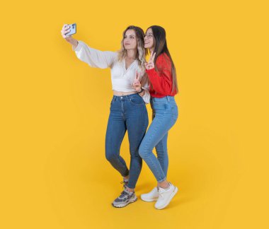 Selfie çekimleri, vücut boyu, güzel, çekici iki genç kadının selfie çekimleri. En iyi arkadaşlar sıradan kot pantolon giyip telefonda fotoğraf çekiyorlar. Zafer işareti sarı renkte..