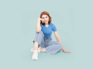 Çok güzel bir vücut, beyaz tenli kızıl saçlı kadın yere oturup gülümsüyor ve kameranın pozitif yüz ifadesi takınıyor. Beyaz spor ayakkabı, mavi kot ve tişört giy. İzole edilmiş mavi arka plan. Boşluğu kopyala
