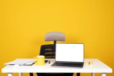 Boş ofis masası, sarı stüdyo geçmişi. Laptop boş beyaz ekran görüntüsüyle maket yapıyor. Kahve fincanı, kağıt belgeler iş malzemeleri ve fotokopi alanı. Çalışanların fikir görüntüsü işe alınıyor.