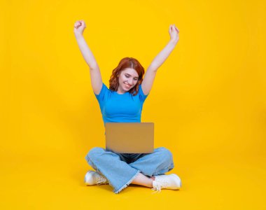 Tüm vücudu mutlu, beyaz, mavi tişört giyen, kot giyen, yerde bacak bacak bacak üstüne atmış, sarı stüdyo arka planında izole edilmiş dizüstü bilgisayar kullanan genç, beyaz bir kadın..