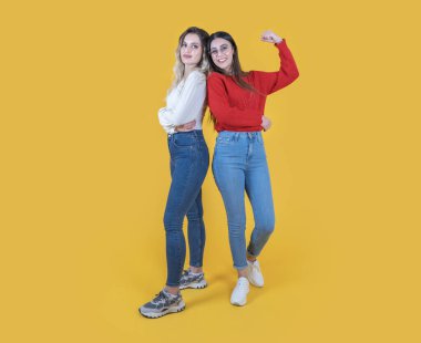 İki kendine güvenen kadın, beyaz tenli güzel iki kadının sarı stüdyo arka planında poz verirken tam vücut görüntüsü. Kutlama jesti, konsept fikir fotoğrafının birincisi. İlk gösteri pazılarını kaldırın..