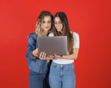 Dizüstü bilgisayar kullanarak, mutlu Kafkasyalı iki genç iş kadını kırmızı stüdyo arka planında dizüstü bilgisayar kullanıyor. Mutlu arkadaşlar gülümseyen bilgisayar ekranına bakıyorum. İş hakkında konuşmak, kopyalama alanı.