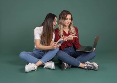 Birlikte dizüstü bilgisayar kullanarak, tüm vücut uzunluğunu kullanarak, iki beyaz kadın dizüstü bilgisayarla zemin bacak üstüne atarak izole edilmiş yeşil stüdyo arkaplanını işaret ediyorlar. Çevrimiçi teknoloji kavramı.