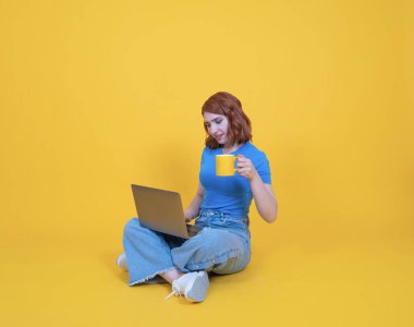 Dizüstü bilgisayar kullanarak, tam vücut ölçülerinde, beyaz kadın dizüstü bilgisayar kullanarak zemin bacak üstüne atarak izole edilmiş sarı stüdyo arka planında kahve içerek, fotokopi çekerek. Uzak konsept çalışıyor.