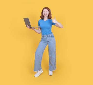 Tüm vücut uzunluğu iyimser, 20 'li yaşlarda güzel bir kadın sarı stüdyo arka planında yalnız poz veriyor. Günlük kot ve mavi tişört giyiyor ve dizüstü bilgisayarını açık tutuyor..