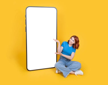 Yeni mobil uygulama sunar, tüm vücudu gören beyaz kadın büyük akıllı telefonun yanında oturur yeni mobil uygulama sunar, telefon sarı arka planda taklit edilir..