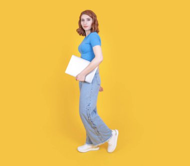 Dizüstü bilgisayarı var, uzun boylu, yan görünüşlü, sarı arka planda, dizüstü bilgisayar taşıyan beyaz tenli, çekici bir kadın. Kadınlar rahat kot pantolon ve mavi tişört giyer. Boşluğu kopyala.