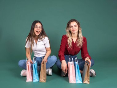 Mutlu tam vücut, iki kadın gündelik kıyafetler giyer. Zemin bacak bacak üstüne atar, yeşil arka planda izole edilmiş kağıt alışveriş torbalarıyla. İndirimli satın alma konsepti fikri imajı, kopyalama alanı.