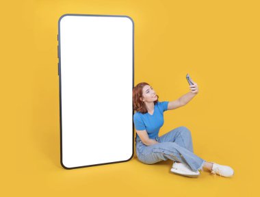 Selfie çekiyor, 20 'li yaşlarda kızıl saçlı kadın rahat kot pantolon ve tişört giyiyor. Büyük cep telefonuna yaslanmış beyaz dokunmatik ekranla taklit ediyor. Sarı arka planda izole edilmiş selfie çekiyor..