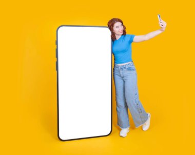 Gülümseyen genç beyaz kadın büyük bir cep telefonunun yanında boş beyaz dokunmatik ekranla selfie çekiyordu. Uygulama çevrimiçi reklam konsepti resmi sunar.