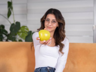 Elinde yeşil elma, gülümseyen mutlu vejetaryen, yeşil elma görünümlü kamera tutan çekici bir kadın. Evdeki fotokopi odasında oturmuş gülümsüyor. İnsanların yaşam tarzı gıda fikri.