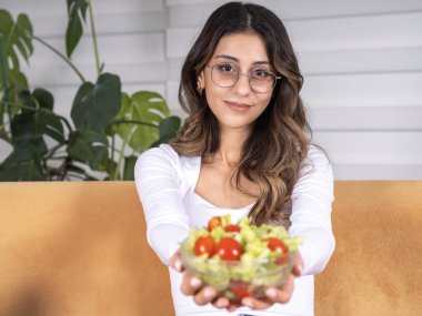 Sağlıklı beslenme, sağlıklı beslenme konsepti imajı için salata dolu bir kâse tutan güzel, genç, fit, beyaz esmer kadın. Evdeki kameraya bakan kanepe. Yeşil salata ve domates.