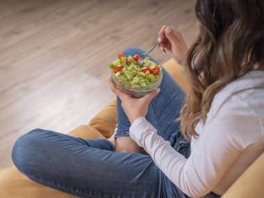 Salata yiyor, omuz hizasında salata yiyen esmer beyaz kadın. Taze yeşil marul ve çatal kullanan dilimlenmiş domates. Evdeki kanepe, sağlıklı yaşam tarzı, diyet yemek konsepti.. 