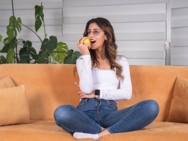 Yeşil elma yiyor, tüm vücudu görüyor, beyaz tenli çekici esmer kadın evde oturup yeşil elma yiyor. Sağlıklı yaşam tarzı kavramı. Taze meyve ısırmak, yan bakmak. Rahat giyin..